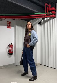 Femme aux longs cheveux foncés debout dans un coin d'un espace industriel, portant un chemisier rayé, un cardigan bleu clair, un jean foncé, des bottes noires, tenant un sac noir.