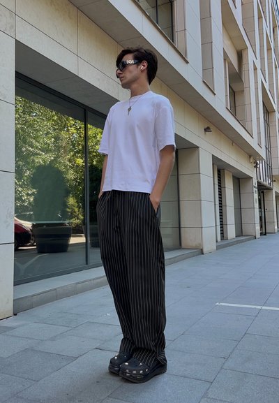 Camiseta blanca de manga corta, pantalones de pierna ancha con rayas negras y zuecos negros con detalles. De pie en una acera con un fondo de edificio de vidrio.