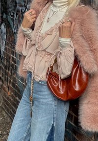 Persona che indossa un cappotto di pelliccia sintetica rosa, una blusa con pizzo, collane a strati, tiene una borsa in pelle marrone, in piedi contro un muro di mattoni coperto di graffiti.