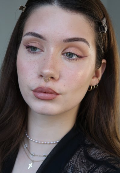 Mujer joven con cabello largo y castaño, maquillaje ligero con delineador de ojos con ala, anillo de septum dorado, múltiples pendientes y collares en capas.