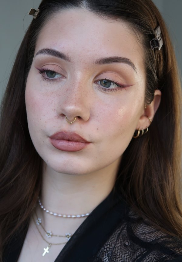 Ung kvinde med langt brunt hår, let makeup med katteøjen eyeliner, guld septumring, flere øreringe og lagdelte halskæder.