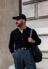 Suéter preto estilo henley com botões, calças cinzentas de cintura alta, cinto de pele preta e um boné preto. A segurar uma grande mala tote preta.