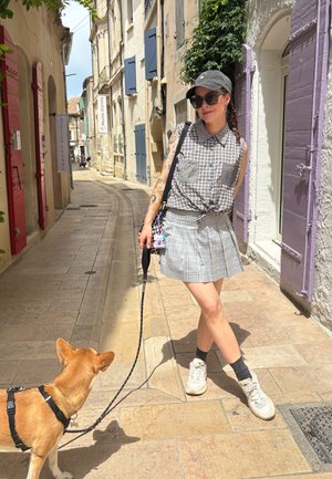 Eine Person in einem karierten ärmellosen Hemd und einem schottischen Rock steht auf einer engen Straße und hält die Leine eines Hundes. Der Hund trägt ein Geschirr.