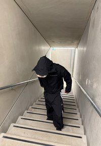 Osoba u crnoj jakni s kapuljačom i hlačama penje se uskim betonskim stubištem s metalnim ogradama s obje strane.