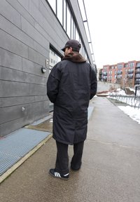 Άνδρας με μακρύ μαύρο παλτό, καφέ φούτερ με κουκούλα, μαύρο καπέλο και μαύρα παπούτσια Adidas, στέκεται σε βρεγμένο πεζοδρόμιο δίπλα σε μοντέρνο κτίριο.