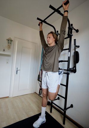 Hombre con prótesis de pierna realizando un ejercicio de dominadas en una barra de gimnasio montada en la pared negra, en una habitación luminosa con suelo de madera.