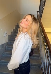 Blondīne, kas valkā baltu džemperi un tumšas džinsas, stāv uz kāpnēm, fonā redzama teksturēta akmens virsma un metāla margas.