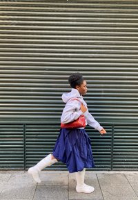 Gaiši pelēks kapučīna džemperis, tumši zils kroku svārks, balti ceļgalu augstie zābaki un sarkana rokas soma. Persona staigā priekšā teksturēta metāla aizvara fonam.