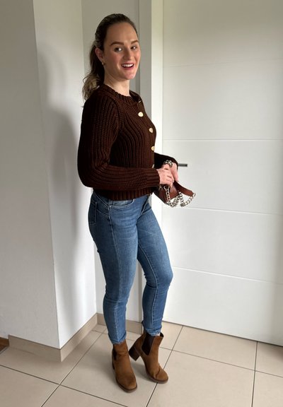 Suéter marrón con canalé y botones dorados, jeans azules y botas de tobillo marrones. El fondo es un interior de color claro. Sosteniendo un bolso pequeño.