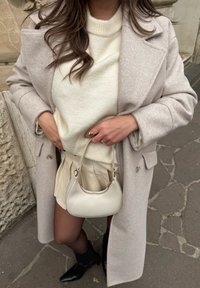 Abrigo de lana gris claro sobre un suéter blanco roto, falda plisada crema y botas negras hasta la rodilla. Sosteniendo un pequeño bolso estructurado crema.