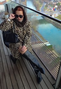 Manteau à imprimé léopard avec boutons rouges, écharpe marron, lunettes de soleil noires surdimensionnées, sac à main noir et bottes noires montant jusqu'aux cuisses, sur un balcon.