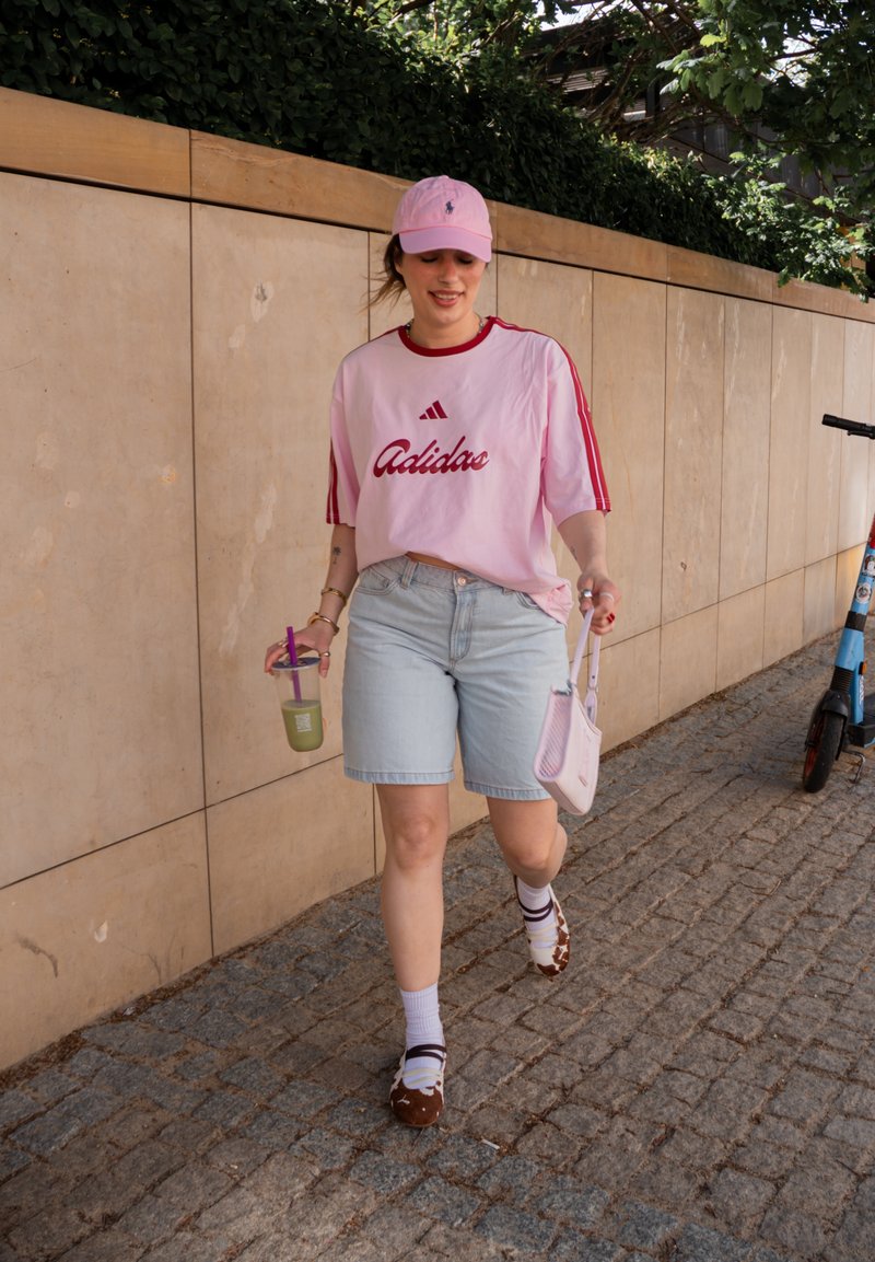 Femme en t-shirt Adidas rose, shorts en jean et casquette rose marchant dans une rue pavée, tenant une boisson et un petit sac, trottinette électrique à proximité.
