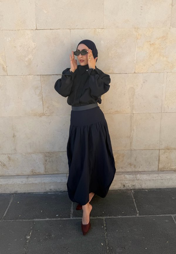 Femme portant un hijab noir, un chemisier noir à manches longues, une longue jupe noire et des talons marron, debout contre un mur en pierre beige en ajustant des lunettes de soleil noires.