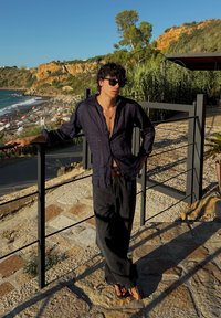 Camisa de lino en azul marino, desabrochada en el cuello; pantalones negros holgados; chanclas; de pie junto a una barandilla negra con vista costera.