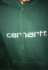 Grøn hættebluse lavet af blødt stof med en frontlomme, snøre i hætten og hvid tekst "carnartt" tværs over brystet.