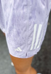 Lys lilla idrettsshorts med våte flekker og hvite Adidas-striper båret på et bein mot en uskarp utendørs bakgrunn.