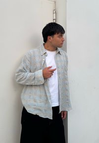 Cămașă în carouri de culoare bleu deschis și bej, cu închidere cu buton, purtată peste un tricou alb, asortată cu pantaloni negri; pe un fundal simplu.