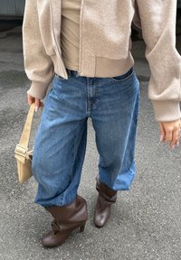 Baggy blå denimjeans, brune hælede støvler med foldet design, og en lys beige cropped sweater med lynlås og ribbede manchetter.