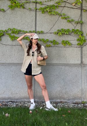 Camisa beige de botones, pantalones cortos negros, zapatillas blancas con acentos verdes, gorra clara, de pie contra una pared gris texturizada con vides verdes.