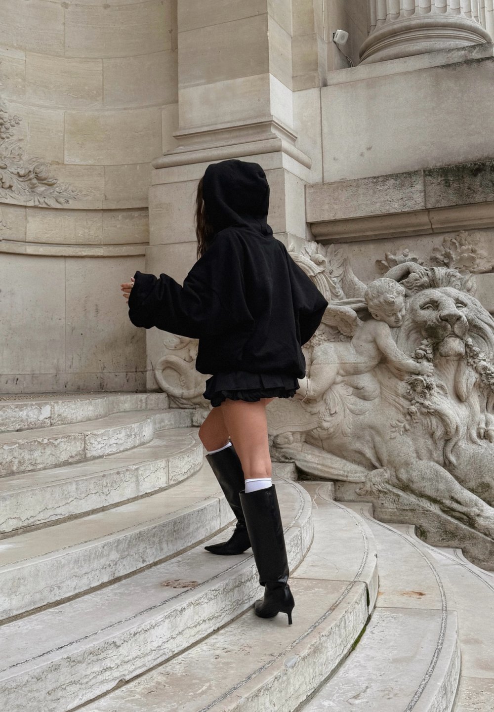 Sweat à capuche noir surdimensionné, jupe noire à volants, bottes noires montantes avec accents blancs, montant des marches en pierre près de sculptures ornées.