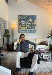 Halli turtleneck, valged püksid ja mustad jalanõud on seljas inimesel, kes istub mustal pouf'il. Taustal on koorevärvi nahksofa, abstraktne seina kunst ja põrandalamp.