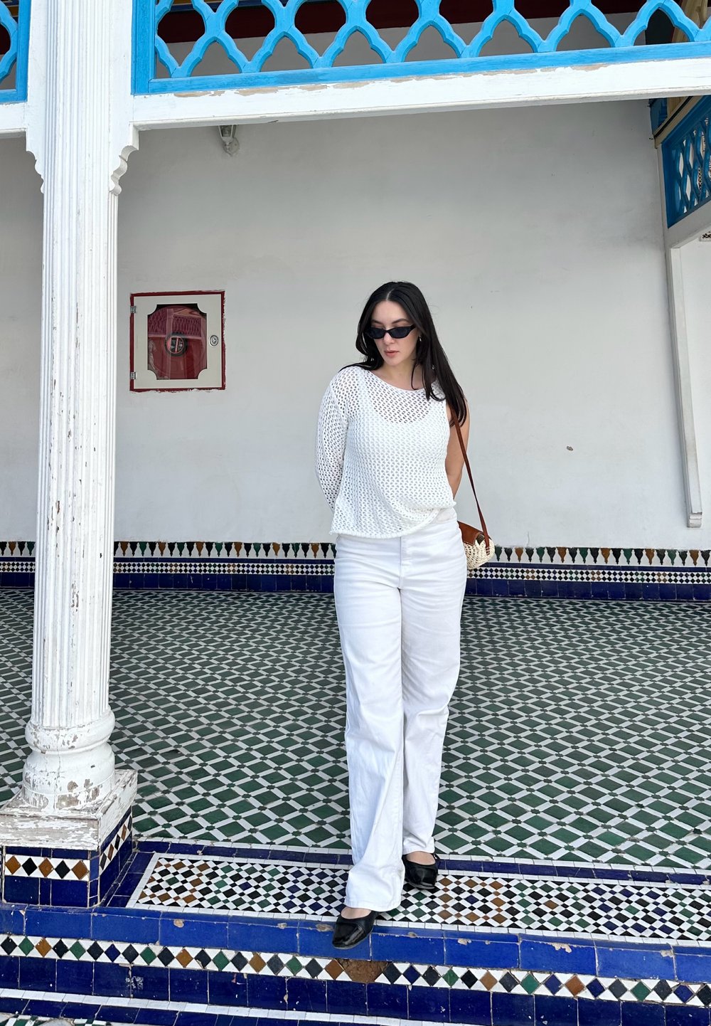 Λευκό πλέγμα μπλούζα μακρυμάνικη, λευκά παντελόνια σε φαρδιά γραμμή, μαύρες παπούτσια με τακούνι και μια ανοιχτή καφέ τσάντα ώμου. Γεωμετρικό πλακάκι στο φόντο.