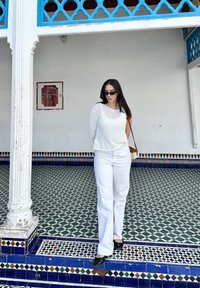 Λευκό πλέγμα μπλούζα μακρυμάνικη, λευκά παντελόνια σε φαρδιά γραμμή, μαύρες παπούτσια με τακούνι και μια ανοιχτή καφέ τσάντα ώμου. Γεωμετρικό πλακάκι στο φόντο.