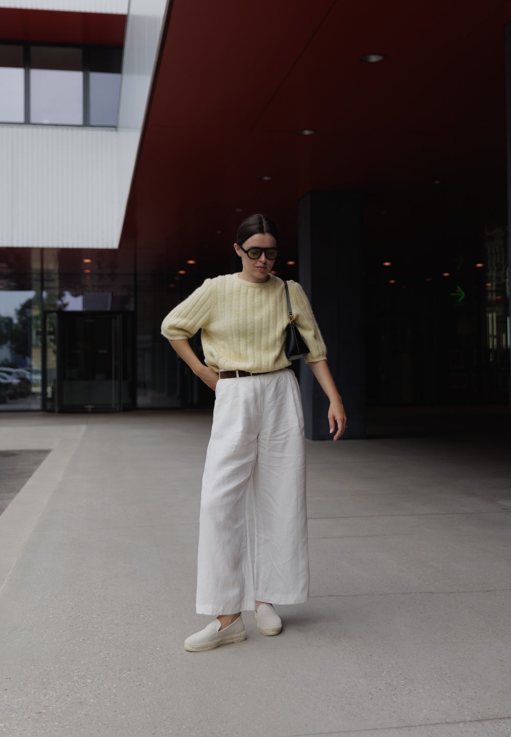 Světle žlutý žebrovaný svetr, volné bílé kalhoty s širokými nohavicemi a béžové slip-on boty. Černá kabelka a převelké černé sluneční brýle.