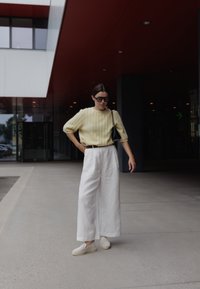 Světle žlutý žebrovaný svetr, volné bílé kalhoty s širokými nohavicemi a béžové slip-on boty. Černá kabelka a převelké černé sluneční brýle.