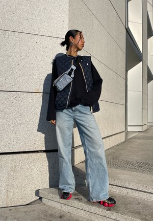 Chaleco acolchado navy, camiseta de manga larga negra, vaqueros de pierna ancha azul claro y zapatillas negras y rojas. Pequeño bolso cruzado gris en el hombro derecho.