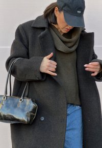 Manteau en laine sombre surdimensionné, pull en maille vert olive avec col bénitier, jean en denim bleu, et petit sac à main en cuir noir avec des poignées courtes.