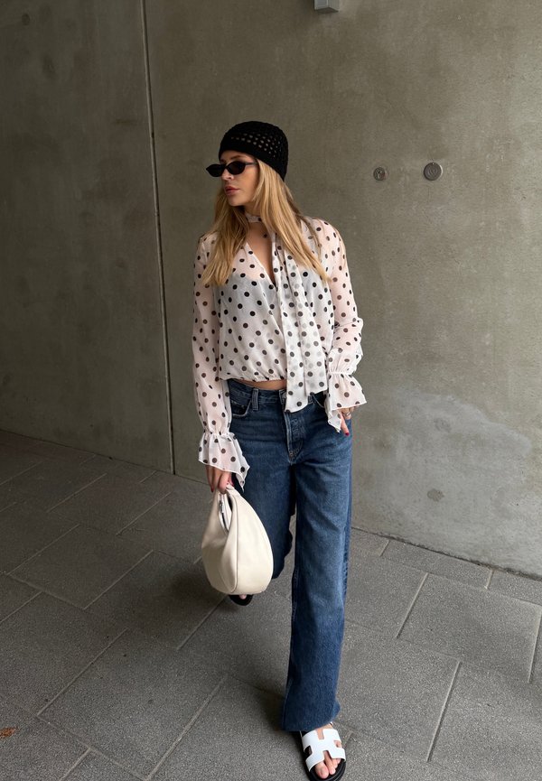 Femme portant un bonnet en tricot noir et des lunettes de soleil, porte un chemisier blanc à pois, un jean bleu, des sandales blanches, et tient un sac à main blanc contre un mur gris.