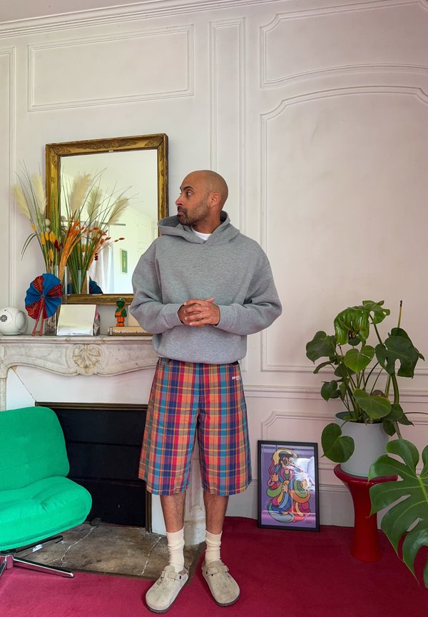 Homme en sweat à capuche gris et short à carreaux se tient dans une pièce décorée avec une chaise verte, des plantes, des œuvres encadrées et un miroir au mur.