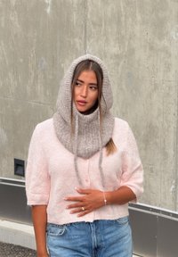 Světle růžový kardigan s krátkým rukávem a knoflíky, nošený přes modré džíny. Obsahuje dvoubarevnou šedou pletenou kapuci se šňůrkami a texturovanou látku.