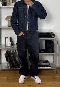 Jachetă din denim închis, cu un design scurt, purtată cu pantaloni negri largi și pantofi sport albi. Tatuaje vizibile, stând într-o cameră cu decor.