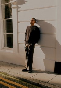 Donker gestructureerd sweater, lichte broek en zwarte schoenen. Staand tegen een witte muur met zonlicht dat schaduwen creëert, wat een casual uitstraling geeft.