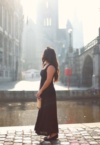 En kvinde i en sort, ærmeløs maxikjole står ved en vandvej og holder en stråhåndtaske. Sollyset skaber en strålende baggrund.