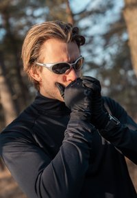 Gants noirs ajustés avec une finition texturée, portés par une personne vêtue d'une chemise noire à manches longues et de lunettes de soleil. Fond de forêt ensoleillée.