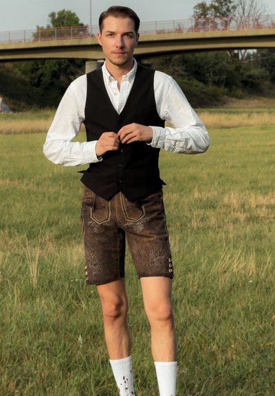 Chaleco negro sobre camisa blanca, pantalones cortos marrones con detalles bordados, calcetines blancos hasta la rodilla, de pie en un campo de hierba.