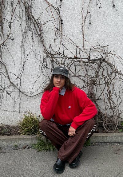 Persona joven con sudadera roja y gorra negra agachada en la acera junto a una pared blanca con ramas secas de vid y pequeñas plantas cerca.