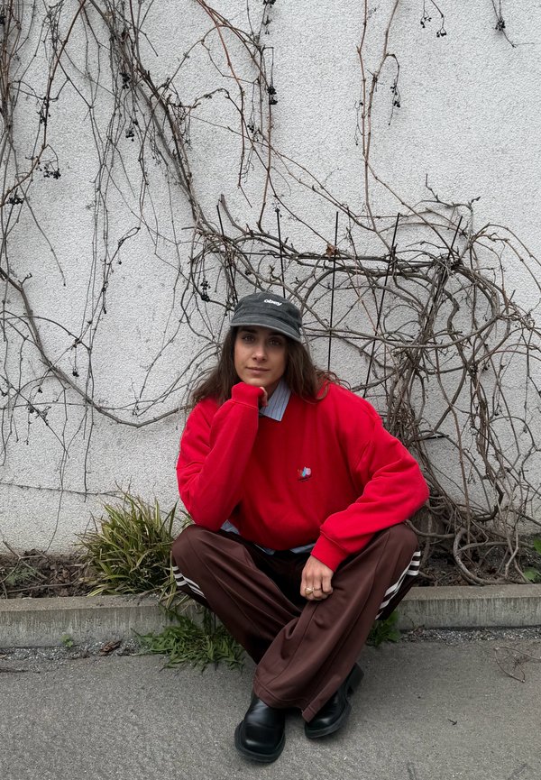 Νέος άνθρωπος με κόκκινο φούτερ και μαύρο καπέλο σκύβει στο πεζοδρόμιο δίπλα σε λευκό τοίχο με ξερά κλαδιά κληματαριάς και μικρά φυτά κοντά.