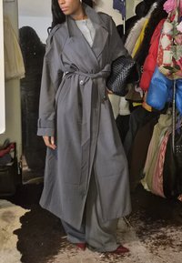 Sieviete, kura valkā garu pelēku mēteļa ar jostu, tur melnu austu maku rokās, stāv pie krāsainiem mēteļiem un somām uz pakaramā.