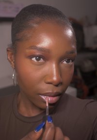 Læbebalsam-applikator holdt tæt på læberne, blank pink glans, lysrefleksion på huden, mørkt hår trukket tilbage, iført en brun top, blå negle synlige.