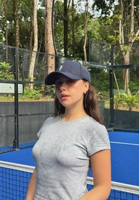 Graues eng anliegendes T-Shirt und marineblaue Kappe getragen von einer Person vor einem blauen Tennisplatz, umgeben von Grünflächen und einem käfigartigen Gehege.