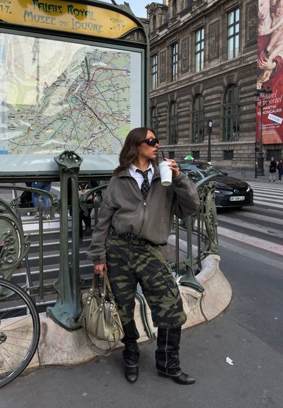 Mujer con gafas de sol, pantalones de camuflaje y chaqueta gris, bebe de una taza cerca de la entrada del metro Palais Royal Musée du Louvre en París.