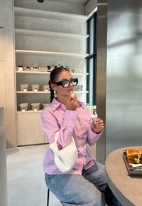 Transparent läppglansapplikator hålls av en person i en rosa randig skjorta. Vit texturerad väska hängande över armen, grå cirkulär bord med rostat bröd.