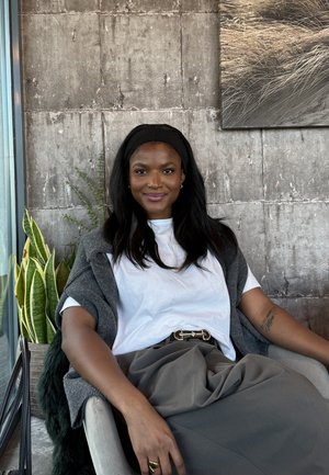 Mujer con cabello largo y negro y diadema, sentada en una silla, vestida con camisa blanca, falda gris y chal, con plantas y una pared de concreto de fondo.