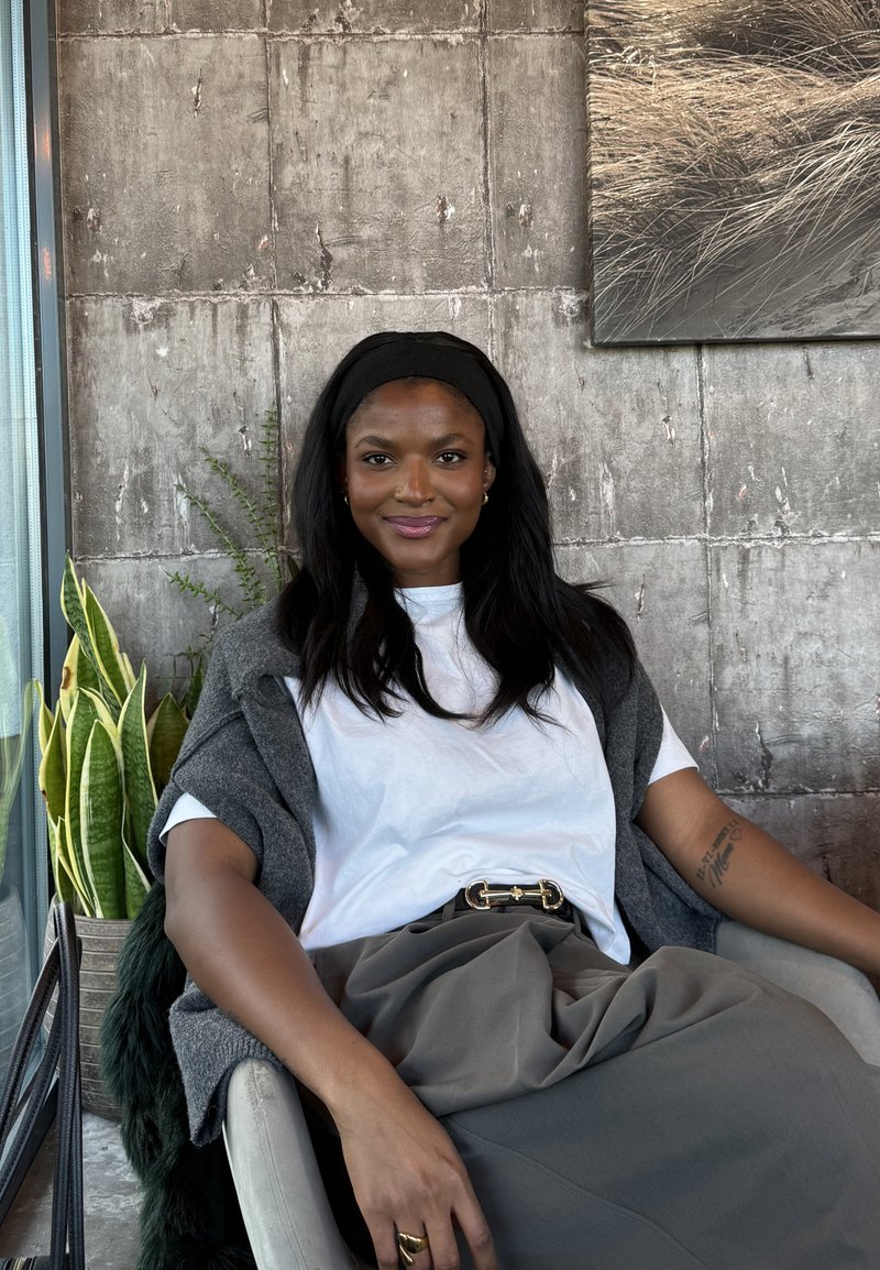 Donna con capelli lunghi neri e fascia in testa seduta su una sedia, indossa camicia bianca, gonna grigia e scialle, con piante e muro di cemento sullo sfondo.