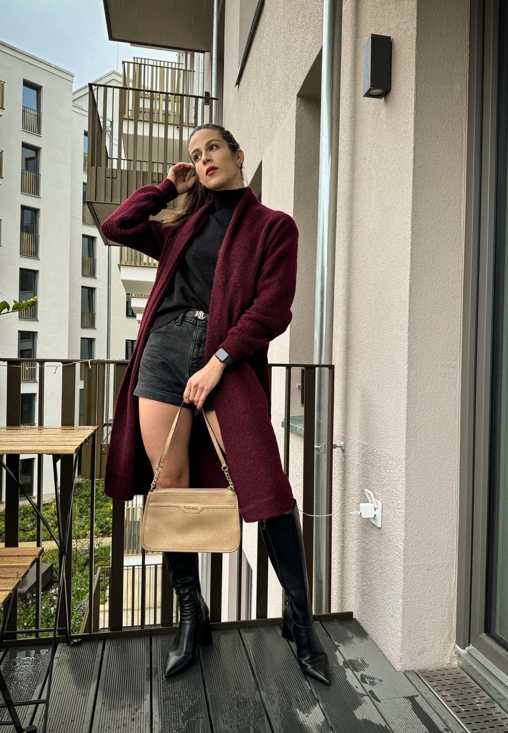 Μπργκούντι πλεχτό μακρύ κάρντιγκαν πάνω από μαύρο ζιβάγκο, σε συνδυασμό με σκούρα σόρτς και μαύρες μπότες μέχρι το γόνατο, κρατώντας μια ανοιχτόχρωμη χρυσή τσάντα.