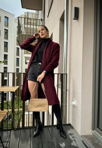 Μπργκούντι πλεχτό μακρύ κάρντιγκαν πάνω από μαύρο ζιβάγκο, σε συνδυασμό με σκούρα σόρτς και μαύρες μπότες μέχρι το γόνατο, κρατώντας μια ανοιχτόχρωμη χρυσή τσάντα.